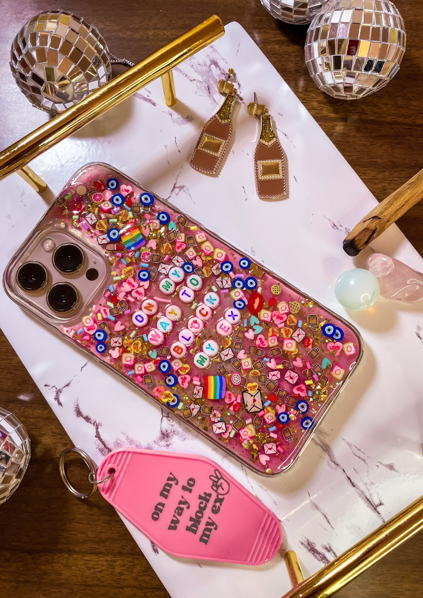 an iPhone case laid flat, decorated with emoji inserts and bracelet beads that read "on my way to block my ex", on a tray surrounded by disco balls